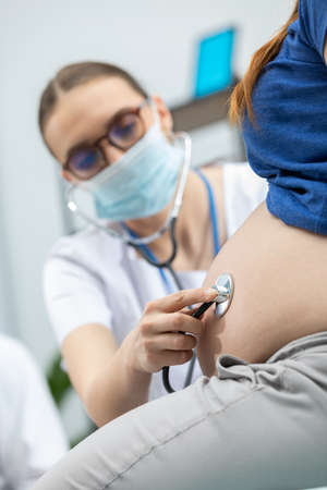 A lady doctor wearing a protective mask examines the abdomen with a stethoscope of a pregnant patient. During a pandemic. Close-up view.の写真素材