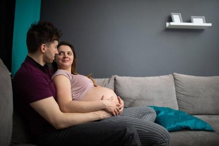 A married couple looks at each other with great joy while sitting on the sofa in the living room. The woman is pregnant.の写真素材