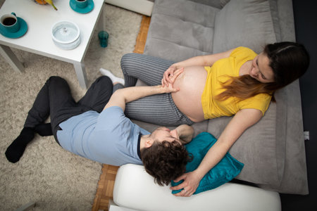 Husband embraces pregnant wifes belly and they smile at each other. Woman in advanced pregnancy. View from above.の素材 [FY310168733297]