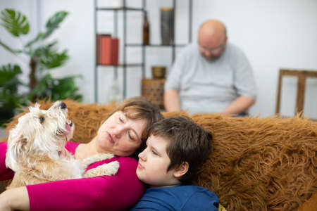 Mother and son are sitting on the sofa and holding a dog in their arms. The father is reading a book.の写真素材