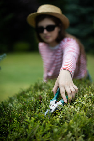 A young woman reaches far with a secateurs to trim a thuja tree branch.