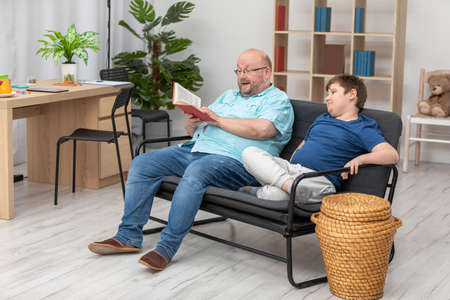A dad is reading an interesting book to his son. They are both sitting on the sofa.