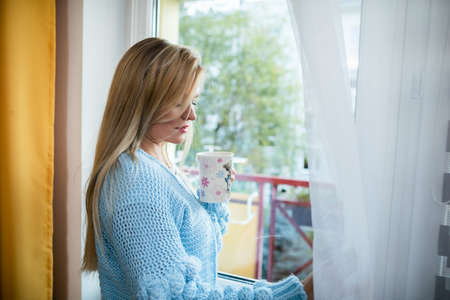 A young woman with a cup in her hand opens the curtain in the window.