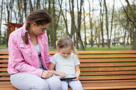 A girl operates a smartphone while sitting on a park bench with her caregiver.の写真素材