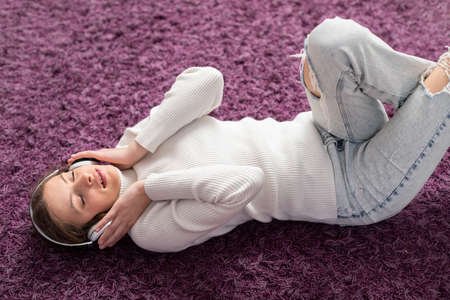 A young girl lies on her back on the carpet and listens to music on headphones.の写真素材