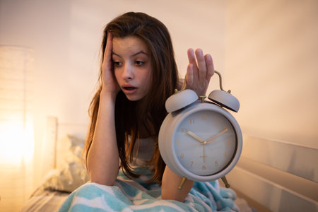 A tired girl holds a bright alarm clock in her hand.の写真素材