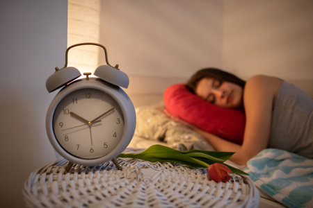 A bedside table with an alarm clock and a tulip stands next to the girl sleeping on her bed.の写真素材
