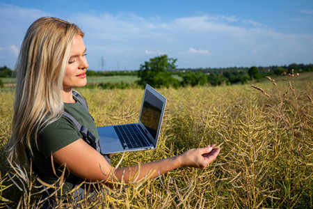 A woman farmer checking the quality of rapeseed, standing at the rapeseed field and holding a laptopの写真素材