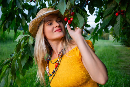 Smiling Blonde Woman in a Mustard Dress Holding Ripe Red Cherries Under a Lush Cherry Tree in a...