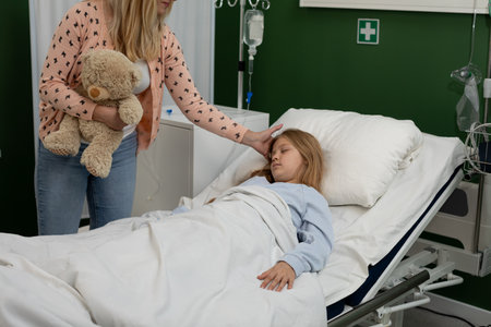 A Child Lying in a Hospital Bed Surrounded by a Caring Adult and a Soft Teddy Bear for Comfortの写真素材