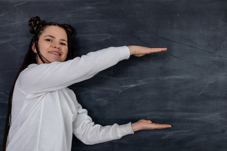 A Cheerful Girl is Gesturing Excitedly in Front of a Black Chalkboard in a Classroom Settingの写真素材