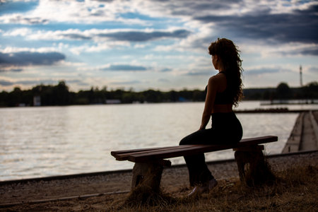 A thoughtful woman is peacefully sitting by the waters edge, contemplating her life at sunsetの写真素材