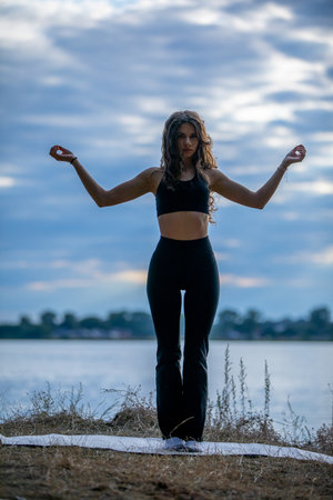 A beautiful silhouette of a woman practicing yoga gracefully by the calming water during duskの写真素材