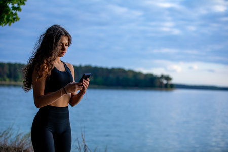 A Young Woman Enjoys the Beauty of Nature by the Tranquil Water While Using Her Smartphoneの写真素材