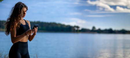 A Young Woman is Relaxing by the Calm Water While Joyfully Using Her Smartphoneの写真素材