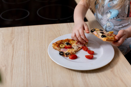 A Happy Child Is Delightfully Enjoying Tasty Pizza with Fresh Ingredients in a Bright Kitchenの写真素材