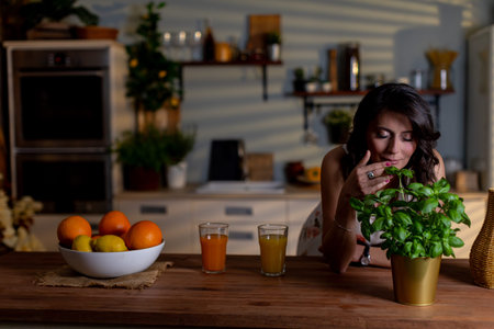 Relaxed woman with herb in kitchen, Serene woman rests in modern kitchen holding fresh basil, Calm female figure lounges at counter with steaming mug and potted plantの写真素材