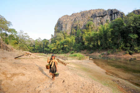 Ban Kong Lo, Laos - 20 january 2012: woman carrying wood logs and basket of salad from the river at the village of Ban Kong Lo on Laosのeditorial素材