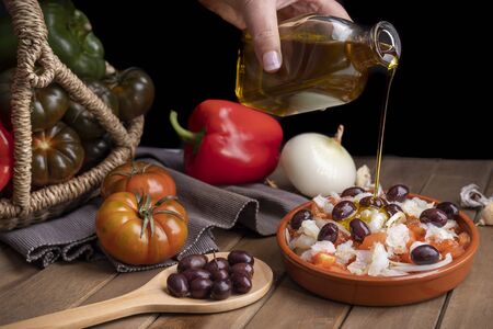Esqueixada in clay casserole with ingredients and wooden spoon with some black olives watering with extra virgin olive oil.の写真素材