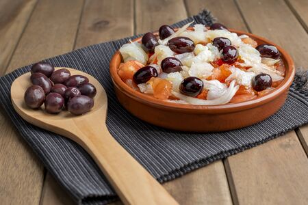 Esqueixada in clay pot and wooden spoon with some black olives on black napkin.の写真素材