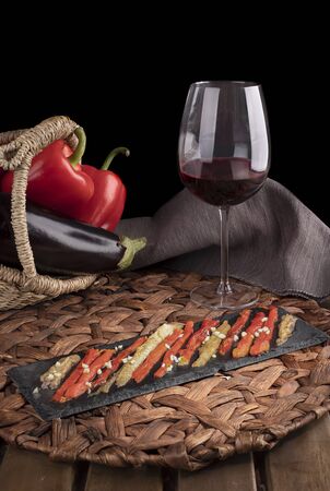 Escalivada with organic food vegetables in wicker basket on rustic wooden table with wine glass. Vertical view.の写真素材