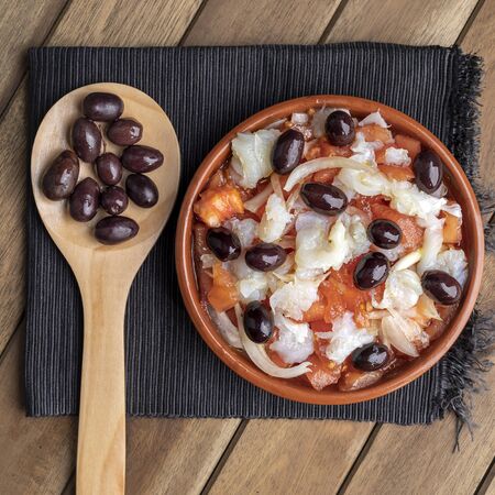 Esqueixada in clay pot and wooden spoon with some black olives on black napkin. Zenith view.の写真素材