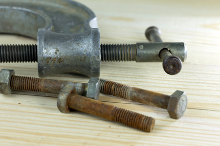 A metal clamp and several bolts are arranged on a wooden surface. The clamp is used for holding objects tightly together. The bolts vary in size and condition.の写真素材