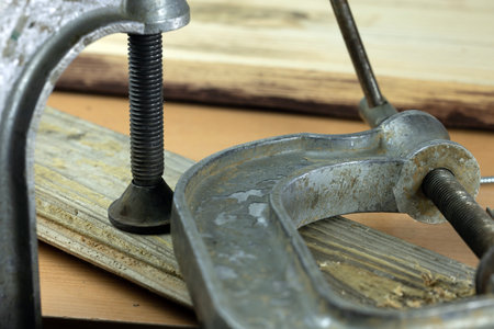 A metal clamp is securing two wooden pieces together on a workbench. The clamp features a threaded screw for tightening and adjusting pressure.の写真素材