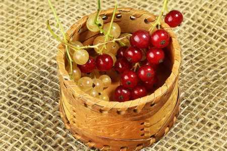 A wooden basket filled with red and green currants on a textured burlap background.