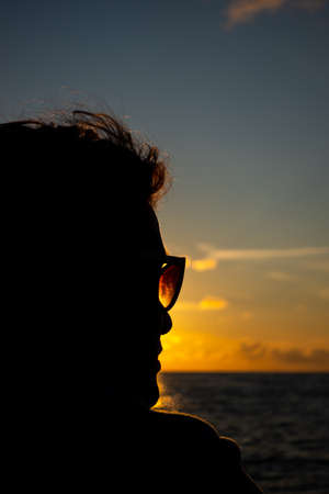 A woman in sunglasses turn her face to the setting sun over the sea. The photo was taken under the atmospheric light of the setting sun.の写真素材
