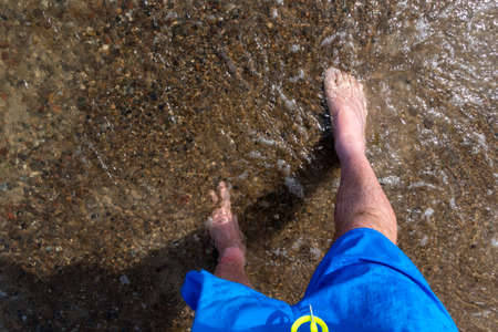 First-person view of the feet of a man standing on the beach, lapped by the gentle waves. Shot was...