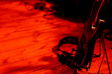 A black electric guitar based on a stand placed on the stage. Subject illuminated by strong red...