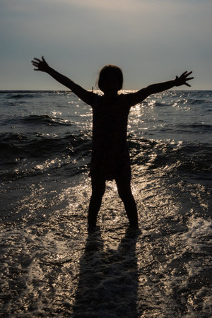 Silhouette of a girl standing in the sea with her arms spread wide. A photo taken against the sun, a figure against the setting sunの写真素材