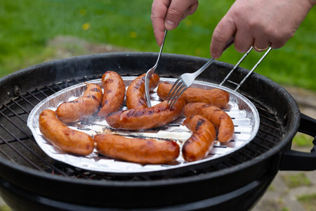 The hands of a person holding two forks change the position of the sausage on the grid of the garden grill. Photo taken in natural lighting conditions.の写真素材
