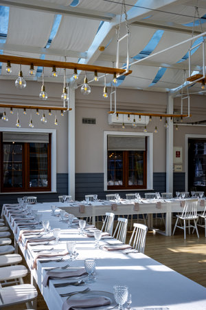 A bright, glass-walled banquet hall with tables covered with white tablecloths. Tableware ready to serve dishes. Photo taken during the day, with sunlight streaming in.の写真素材