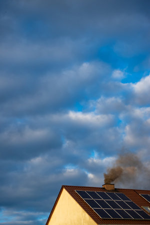 Black smoke from the chimney of a single-family home. Roof covered with solar panels. Roof against a blue, slightly cloudy sky.の写真素材