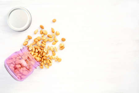 Roasted organic unsalted peanuts spilling from pink jar on rustic white wood background. Shot from...