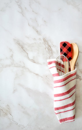 Spatula,wooden spoon,and whisk wrapped in red and white tea towel on marble background in flat lay format. In vertical composition with room for text.の写真素材