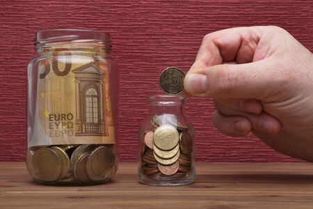 Hand holds euro coins and bill on the burgundy red background in a money box, isolated on the tableの写真素材