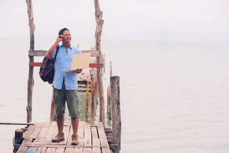 Asian male tourist standing at old wooden bridge using computerの写真素材