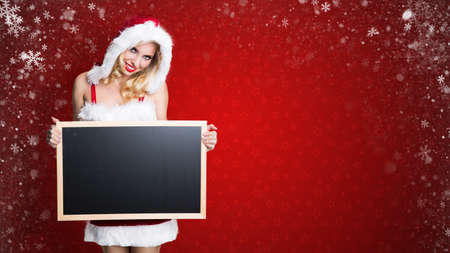 attractive miss santa with an empty blackboard in front of a christmas backgroundの写真素材