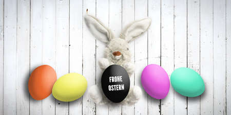 Stuffed bunny with message Hapy Easter in German and many easter eggs on white wooden background