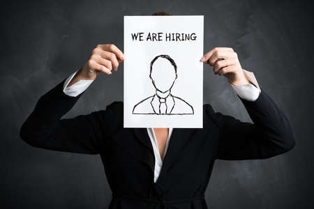 businesswoman holding a sign with the message "we are hiring"  in front of a blackboardの写真素材