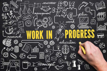 hand is drawing business doodles and the text "work in progress" on a blackboardの写真素材