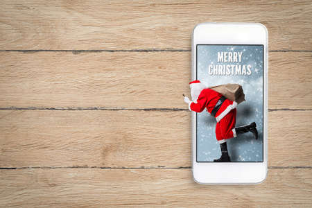 Santa Claus running out of a smartphone and Merry Christmas message on wooden background