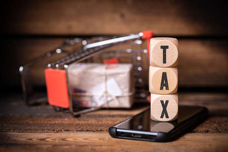 shopping cart laying on the side and cubes with the word TAX on a smartphone on wooden background