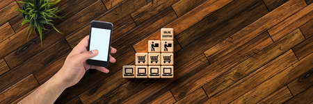 hand with smartphone and cubes with message ONLINE SHOPPING and shopping icons on wooden backgroundの写真素材