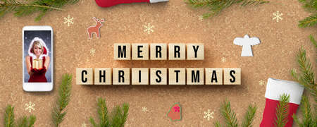 cubes with message MERRY CHRISTMAS besides a smartphone with a smiling miss santa and christmas decoration on cork backgroundの写真素材