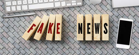 wooden blocks as domino blocks with the message FAKE NEWS surrounded by a smartphone and computer keyboard on stone pavement backgroundの写真素材