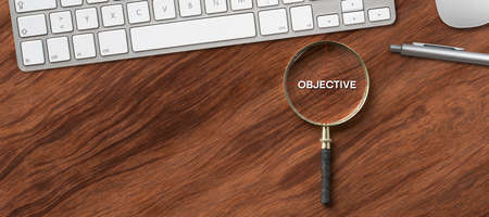 Magnifying glass with text - Objective - on target icon on a wooden desk in an office with computer...